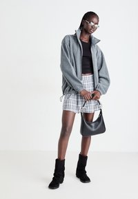 Graue Fleecepullover-Jacke, schwarzes Cropped-Top, karierte Shorts, schwarze Wildlederstiefel und eine kleine schwarze Handtasche. Das Model posiert in einem minimalistischen Setting.