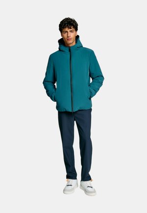 Giacca turchese con zip, cappuccio, maniche lunghe e tasche laterali, abbinata a pantaloni blu navy e scarpe da ginnastica bianche. Tessuto liscio e vestibilità rilassata.