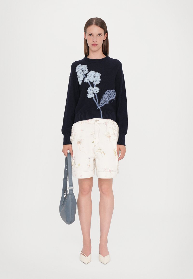 Pullover en maille marine avec broderie florale, short clair jusqu'aux genoux avec un motif floral, associé à un sac à main gris et des chaussures beiges.