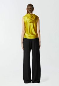 Top giallo in satin senza maniche con cappuccio, abbinato a pantaloni neri a gamba larga. Il top ha una superficie liscia e un orlo arrotondato.