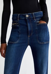 Jeans in denim blu scuro con vita alta, dotati di dettagli in metallo dorato e tasche anteriori. Il tessuto presenta una trama liscia con cuciture visibili.