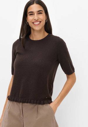 Kvinde med langt mørkt hår iført en mørkebrun kortærmet sweater og beige bukser, smilende med den ene hånd i lommen.