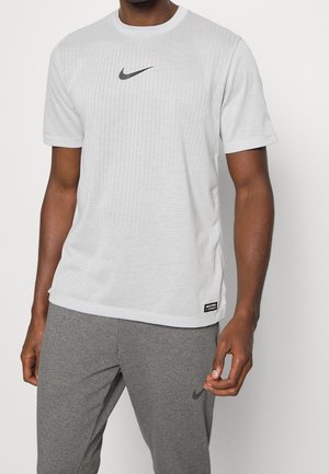 Homme portant un t-shirt blanc à manches courtes Nike et un pantalon de survêtement gris Nike, debout devant un fond blanc uni.