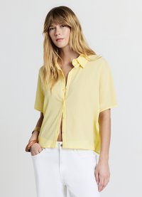 Chemise boutonnée à manches courtes jaune en tissu léger, avec une coupe décontractée et une surface texturée, associée à un pantalon blanc.