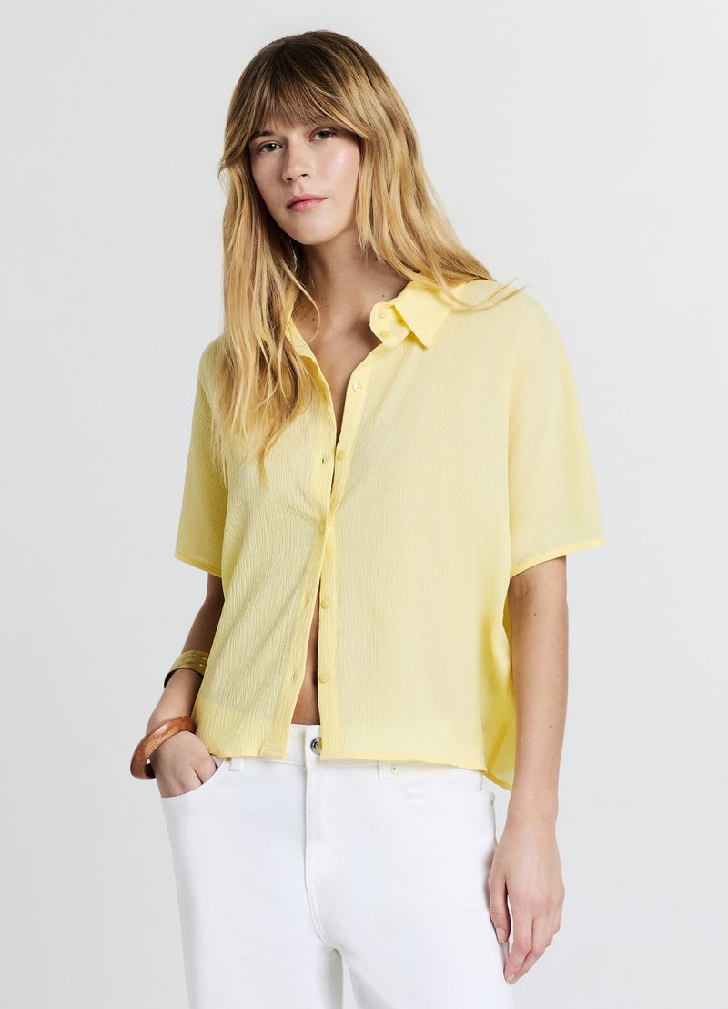Chemise boutonnée à manches courtes jaune en tissu léger, avec une coupe décontractée et une surface texturée, associée à un pantalon blanc.