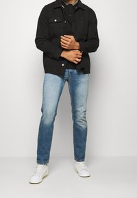 Czarna dżinsowa kurtka z dwoma przednimi kieszeniami, noszona na czarnej koszulce polo. Jasnoniebieskie slim-fit dżinsy i białe trampki dopełniają strój.