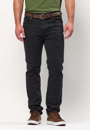 Férfi fekete farmerben, zöld ingben, amit betűrt, barna öv és barna sportcipő, karjai lazán oldalt.