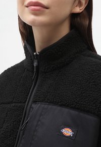 Jacket in fleece nero con colletto alto, tessuto testurizzato e un pannello nero a contrasto nella parte inferiore. Presenta un patch del logo Dickies.