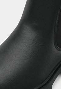 Tuvumā redzams melnā ādas Čelsijas zābaks, kurā redzama gluda tekstūra, elastīga sānu paneļa un izturīga zoles mala.