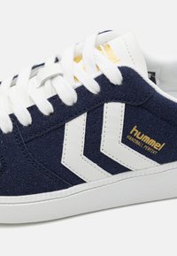 Baskets en daim bleu marine avec des bandes chevron blanches, des lacets blancs et un marquage doré "hummel HANDBALL PERFEKT" sur le côté.