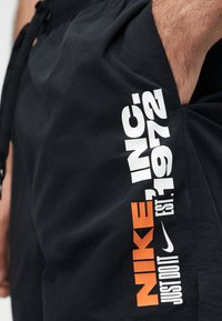 Κοντινό πλάνο μαύρων σορτς Nike με πορτοκαλί και λευκή επιγραφή "NIKE, EST. 1972, JUST DO IT" και λογότυπο swoosh στην περιοχή της πλαϊνής τσέπης.