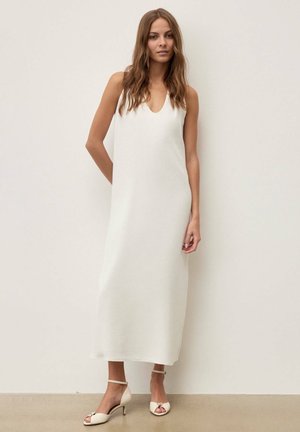 Kvinde med langt brunt hår iført en ærmeløs hvid kjole, der går til anklerne, og hvide sandaler med lav hæl, stående op ad en ensfarvet væg.