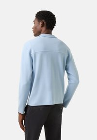 Lyseblå langærmet pullover med krave, glat tekstur og lige kant, kombineret med mørke bukser med baglommer.