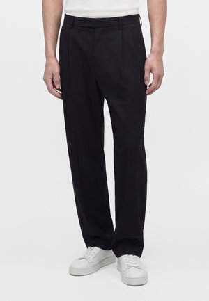 Homme portant un pantalon noir à plis et des baskets blanches, debout devant un fond clair uni.