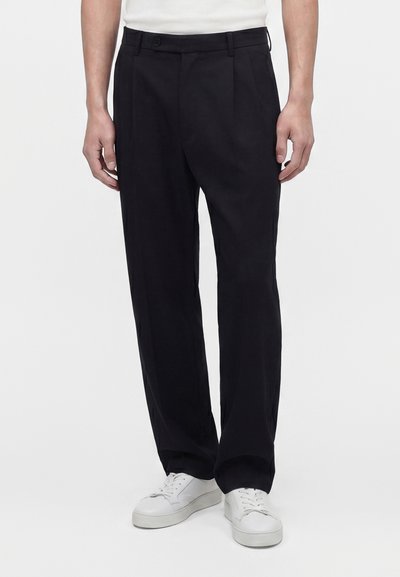 Homme portant un pantalon noir à plis et des baskets blanches, debout devant un fond clair uni.