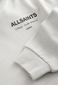 Sudadera gris claro con puños acanalados, que presenta el nombre de la marca "ALLSAINTS" impreso en negro junto con texto en japonés, ubicado en la parte delantera.