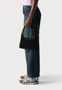 Una persona con jeans blu e sneakers bianchi e marroni tiene una borsa in pelle nera con borchie metalliche al suo fianco.