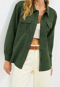 Camicia verde con bottoni, realizzata in tessuto morbido, dotata di due tasche sul petto, vestibilità casual e maniche lunghe, abbinata a pantaloni chiari.