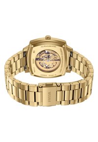 Montre en acier inoxydable doré avec un boîtier carré, un fond transparent et un bracelet à maillons. Présente des détails gravés et une fermeture sécurisée.