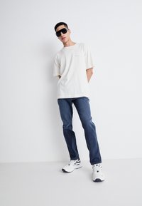 Camiseta de algodón beige, corte relajado, con mangas cortas. Jeans de mezclilla azul, corte recto. Zapatillas blancas con acentos en negro y azul marino.