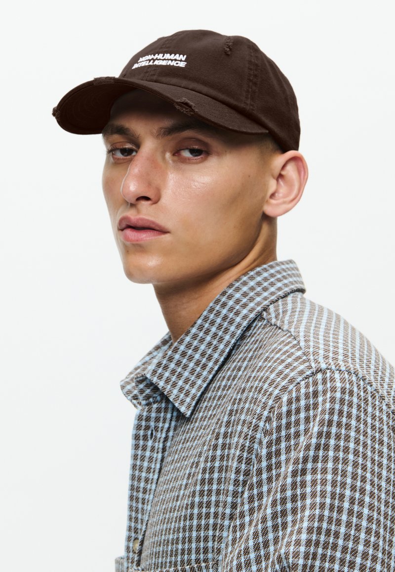 Cappello di cotone marrone con visiera curva e testo ricamato bianco, abbinato a una camicia a quadri blu e marrone con colletto.