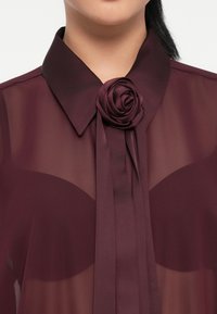 Burgundijska prozorna bluza s strukturiranim ovratnikom i dekorativnim akcentom ruže. Sadrži duge, lepršave kravate koje elegantno vise s ovratnika.