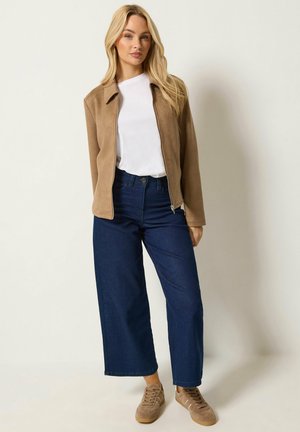 Mujer de cabello largo y rubio lleva una chaqueta marrón con cremallera sobre una camisa blanca, vaqueros de pernera ancha azul oscuro y zapatillas marrones, de pie frente a un fondo liso.