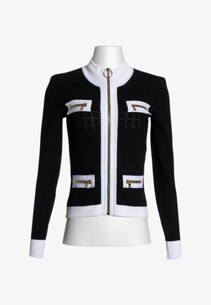 Giacca nera aderente in maglia con rifiniture bianche, cerniera frontale dorata e quattro tasche decorative con zip su petto e vita.