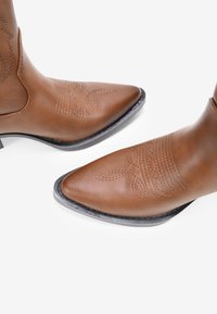 Stivali da cowboy in pelle marrone con punte appuntite, cuciture decorative sulla parte superiore e un tacco basso e impilato. Superficie testurizzata e design elegante.