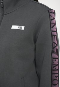 Felpa grigia con zip, caratterizzata da un alto colletto, tessuto testurizzato e una striscia di branding viola prominente lungo il braccio sinistro.