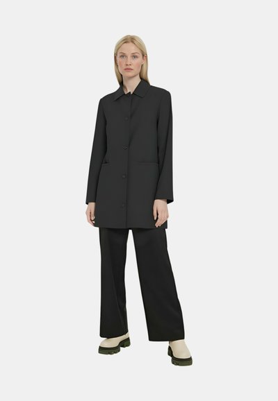 Cappotto nero con chiusura a bottoni, colletto e due tasche. Abbinato a pantaloni neri a gamba larga e stivali alla caviglia color crema con suole verdi.