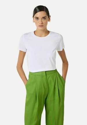 Donna con capelli scuri indossa una camicia bianca a maniche corte infilata in pantaloni larghi verde brillante a vita alta, mani nelle tasche.