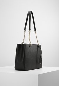 Sac fourre-tout noir en matière synthétique texturée, doté de doubles anses (cuir et chaîne), forme rectangulaire, et une petite étiquette attachée.