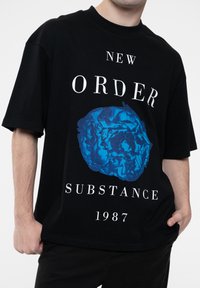 Black cotton t-shirt featuring a blue graphic design of a crumpled object, text "NEW ORDER SUBSTANCE 1987" in bold white lettering.