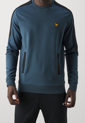 Mand iført blå langærmet sweatshirt med sorte striber på skuldre og lommer, kombineret med sorte Nike bukser.
