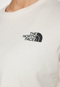 Primo piano di una camicia beige chiaro con il logo nero "The North Face" stampato sulla parte superiore del petto.