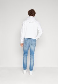 Fehér kapucnis pulóver hosszú ujjakkal, világoskék, slim-fit farmernadrággal és fehér sportcipővel párosítva, hátulról nézve.