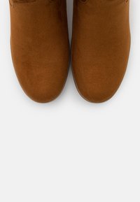 Brązowe zamszowe botki za kostkę z okrągłym noskiem i płaską podeszwą, o gładkiej fakturze i minimalistycznym designie. Brak widocznych akcentów dekoracyjnych.