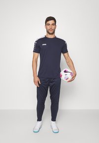 Maglietta sportiva della marina con maniche corte, dettagli bianchi sulle spalle, pantaloni neri e calzature bianche, che tiene un pallone da calcio con dettagli rosa.