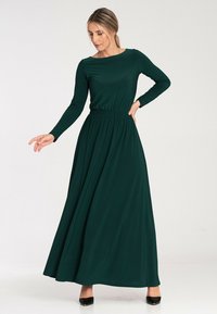 Długa, ciemnozielona sukienka uszyta z gładkiego materiału, z długimi rękawami, dopasowaną talią z gumką i zwiewną spódnicą sięgającą do podłogi.