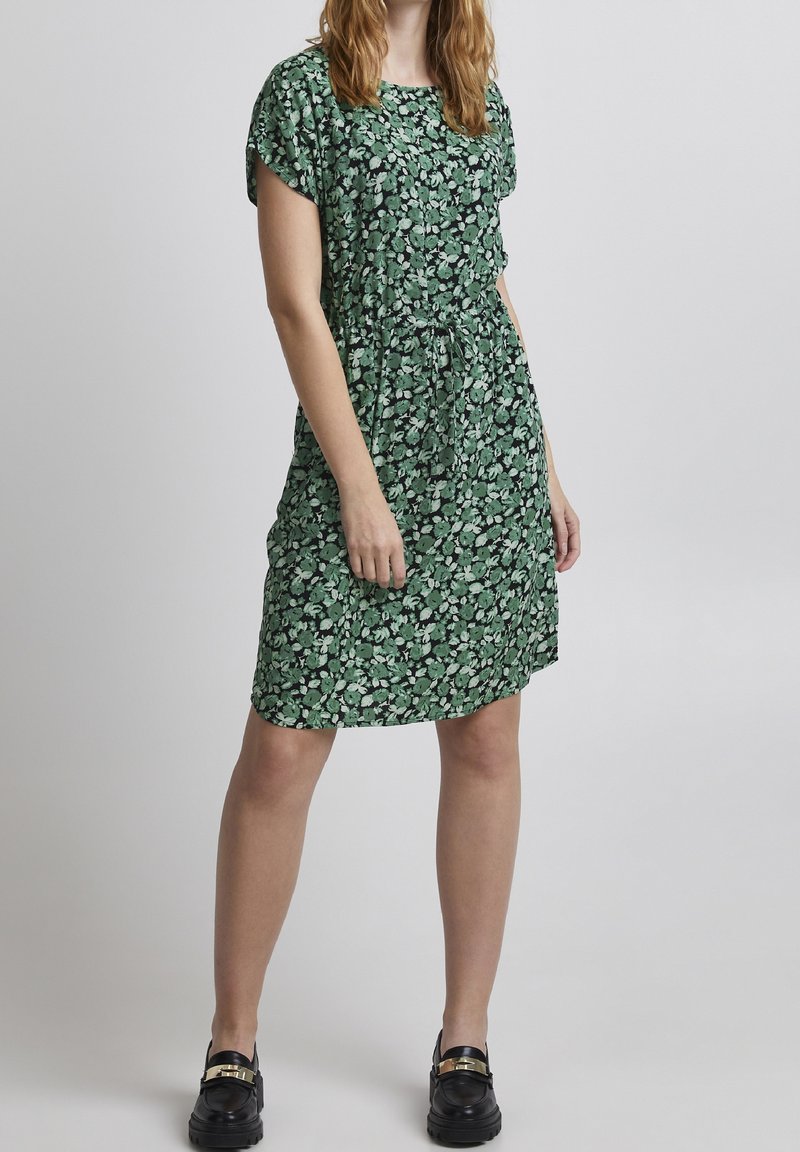 Femme portant une robe verte à fleurs jusqu'aux genoux, à manches courtes, et des mocassins noirs avec un détail de boucle dorée, debout devant un fond blanc.