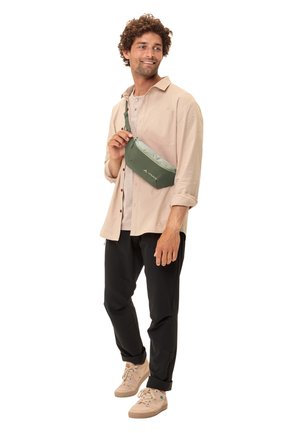 Sac bandoulière vert avec une texture lisse, doté d'une fermeture zippée et d'un logo. Porté sur une chemise claire et un pantalon noir, tenu par un modèle.