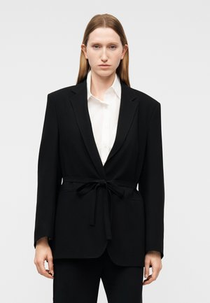 Femme aux cheveux bruns clairs et raides, portant un blazer noir cintré avec ceinture sur une chemise blanche à col, se tenant devant un fond clair uni.