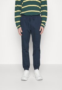 Navyblå joggerbyxor med elastiska muddar och dragsko i midjan, i kombination med en grönrandig långärmad tröja och vita sneakers.