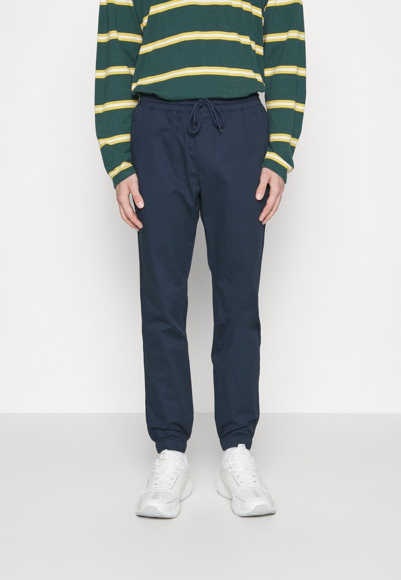 Navyblå joggerbyxor med elastiska muddar och dragsko i midjan, i kombination med en grönrandig långärmad tröja och vita sneakers.