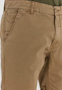 Primo piano di pantaloni casual beige che mostrano la tasca anteriore, i passanti per la cintura, la chiusura con bottone e i dettagli della cucitura.