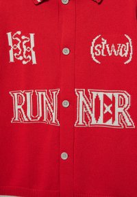 Cardigan en tricot rouge à boutons avec un texte brodé en blanc "RUNNER" et des symboles décoratifs brodés au-dessus du texte.