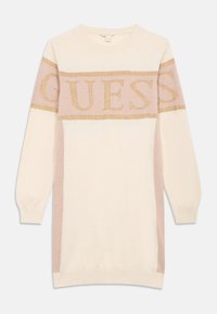 Pletené svetrové šaty v krémové barvě s bledě růžovými bočními panely. Na hrudi se nachází zlatý třpytivý logotyp "GUESS". Dlouhé rukávy, kulatý výstřih.
