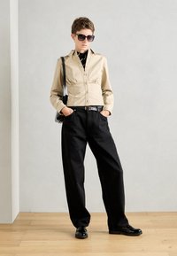 Beige Hemd mit Knopfverschluss und geraffter Taille, kombiniert mit lockeren schwarzen Hosen und glänzenden Schuhen. Das Model trägt Sonnenbrillen und eine Umhängetasche.