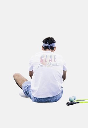 T-shirt blanc avec l'inscription "CLAE Los Angeles" en texte coloré, associé à un short bleu clair. Raquette de tennis et balles sur la droite.